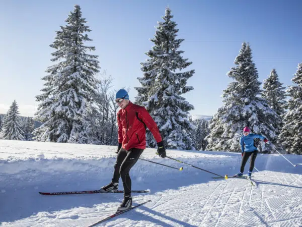 Ihr seht 2 Personen beim Langlaufen auf der Loipe Herzogenhorn mit winterlicher Landschaft im Hintergrund.