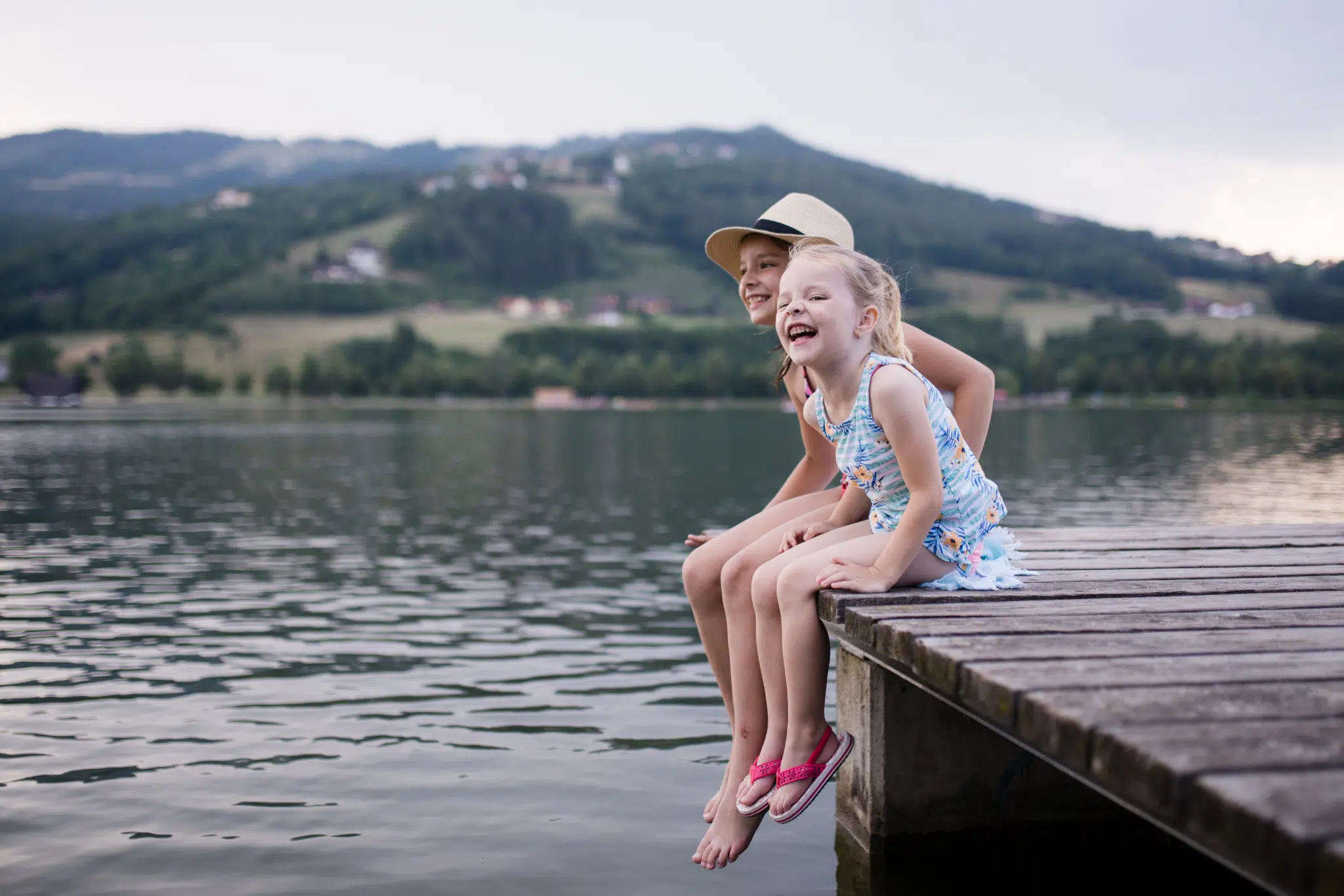 Ihr seht zwei lachende Mädchen auf einem Steg am Stubenbergsee im Sommer. JUFA Hotels bietet tollen Sommerurlaub an schönen Seen für die ganze Familie.