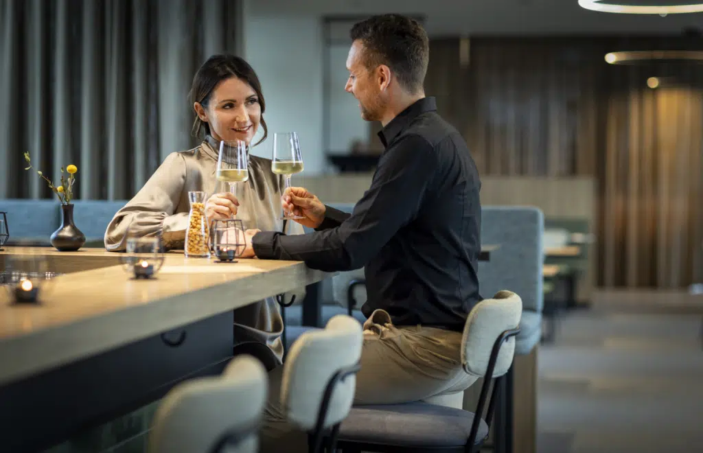 Ihr seht ein romantisches Pärchen bestehend aus Mann und Frau, die gerade mit einem guten Glas Weißwein aus dem Steirischen Thermen- & Vulkanland an der Bar im Café & Restaurant Wandelhalle im JUFA Hotel Bad Radkersburg anstoßen.