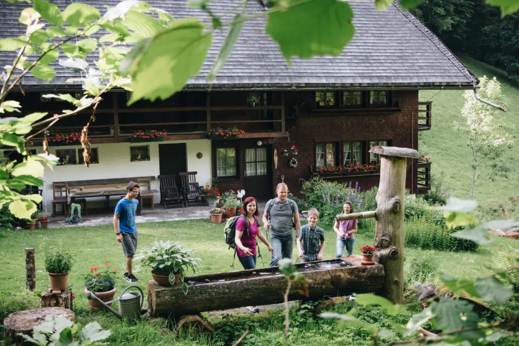 Ihr seht eine Familie, die sich auf einer Alm an einem Brunnen erfrischt und abkühlt. JUFA Hotels bietet Ihnen den Ort für erlebnisreichen Natururlaub für die ganze Familie.