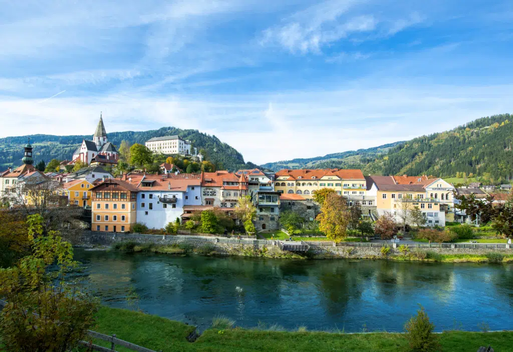 Ihr seht ein Bild von der Mur, dahinter die historische Altstadt Murau mit Blick auf das Schloss Murau und die umliegende Bergwelt.