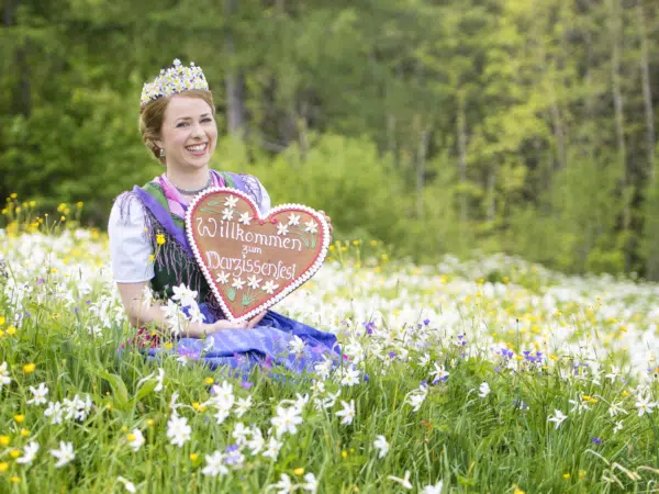 Ihr seht die Narzissenkönigin mit einem Lebkuchenherz in einer Blumenwiese. JUFA Hotels bietet kinderfreundlichen und erlebnisreichen Urlaub für die ganze Familie.