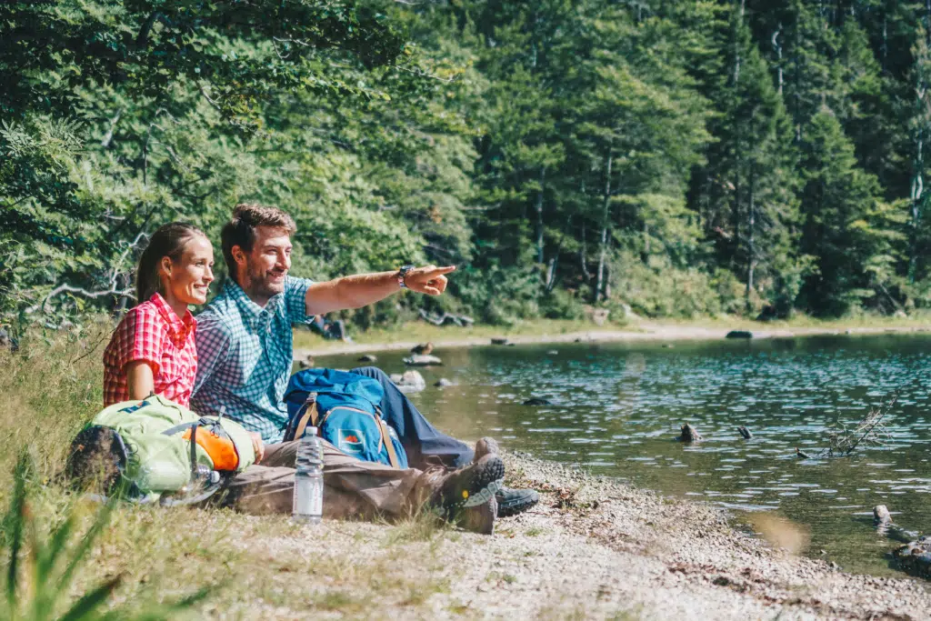 Ihr seht ein Paar, das während einer Wanderung im Schwarzwald eine Pause am wunderschönen Feldsee macht. JUFA Hotels bietet Ihnen den Ort für erlebnisreichen Natururlaub für die ganze Familie.