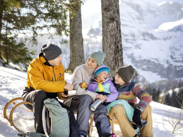 Ihr seht eine Familie mit Vater, Mutter, Tochter und Sohn mit einer Jausenbox und Thermosflasche auf einer Rodel in einer verschneiten, sonnigen Winterlandschaft mit Bergen im Hintergrund.