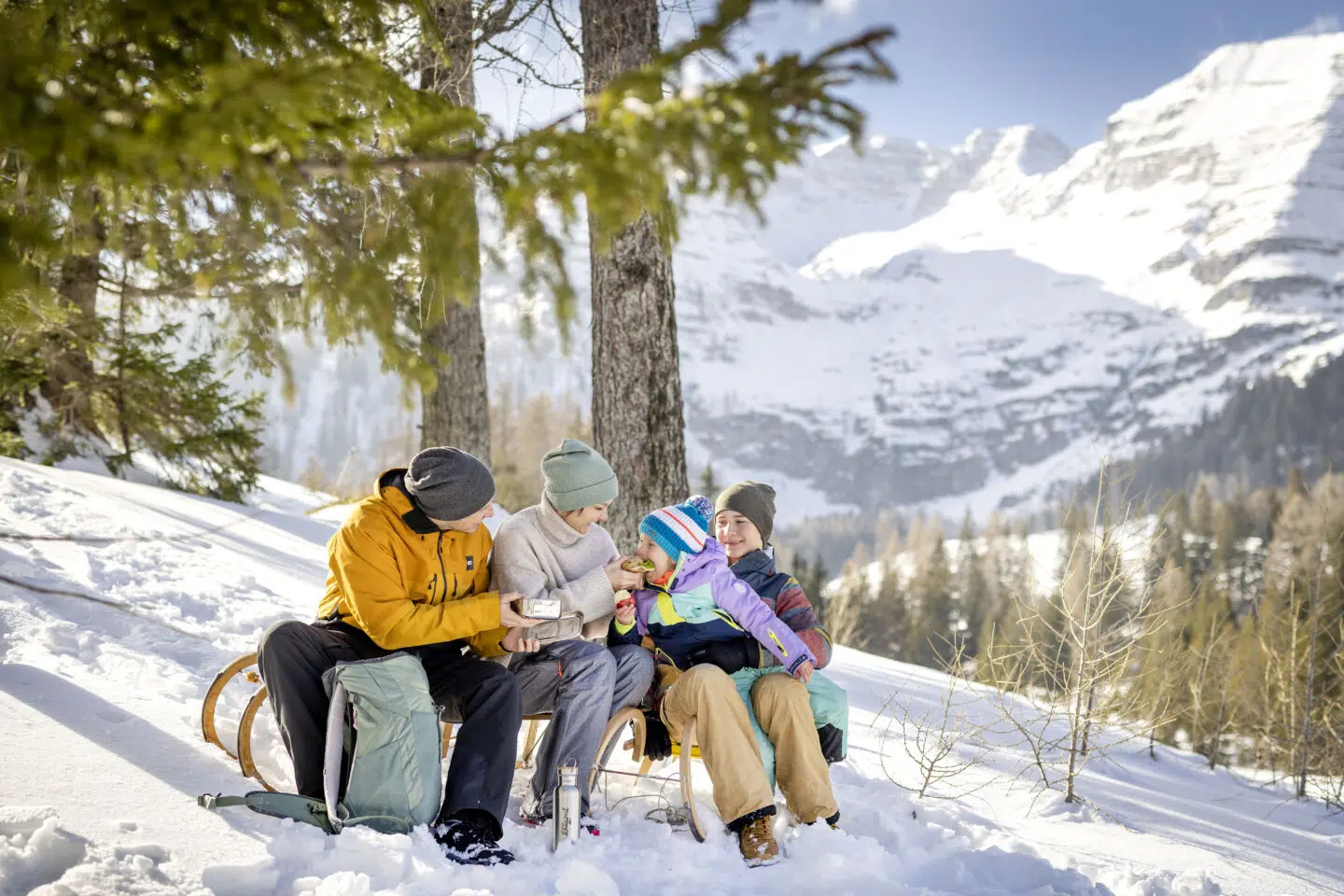 Ihr seht eine Familie mit Vater, Mutter, Tochter und Sohn mit einer Jausenbox und Thermosflasche auf einer Rodel in einer verschneiten, sonnigen Winterlandschaft mit Bergen im Hintergrund.