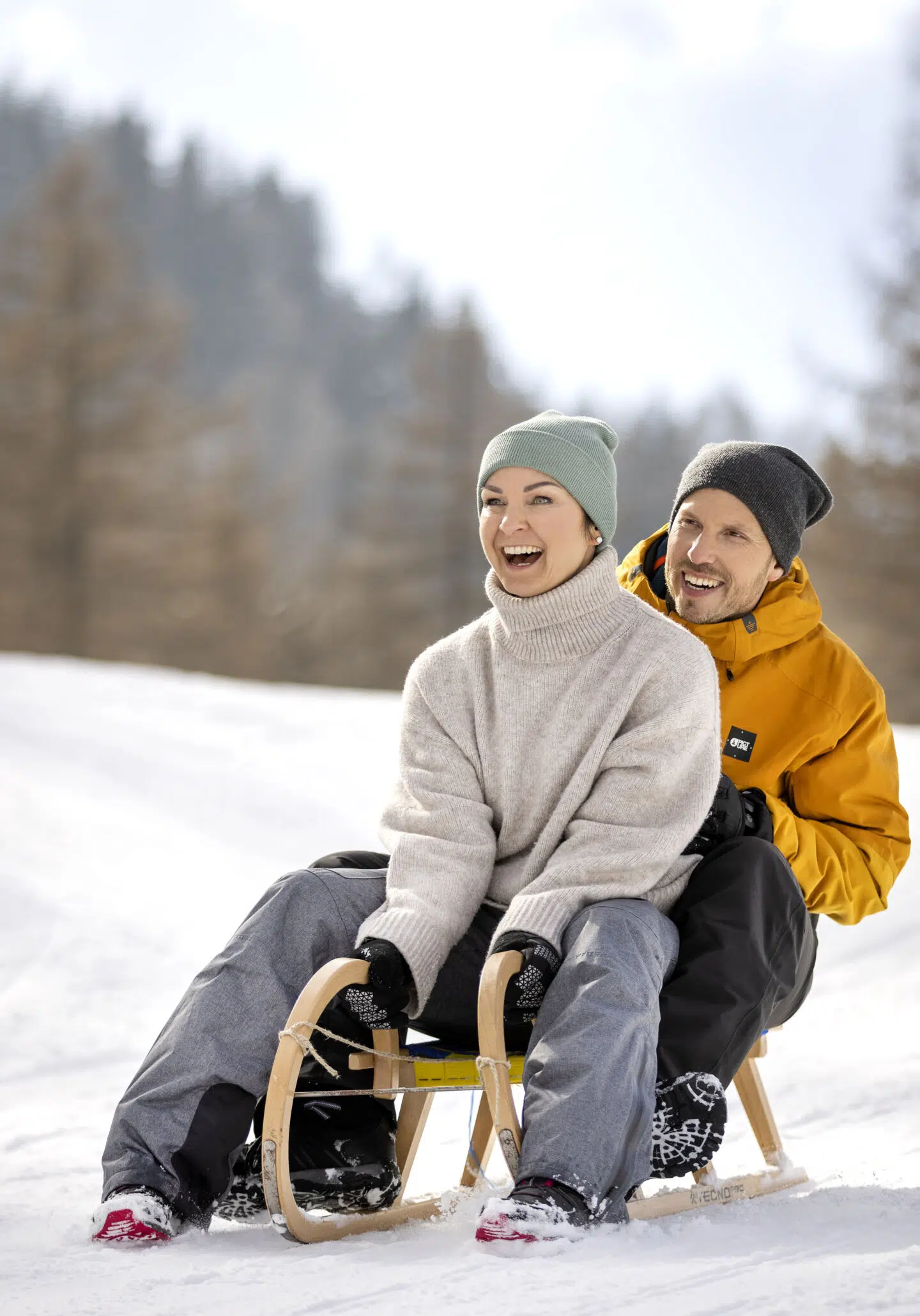 Sie sehen einen Mann und eine Frau, lachend, bei einer Fahrt mit einer Rodel im Schnee.