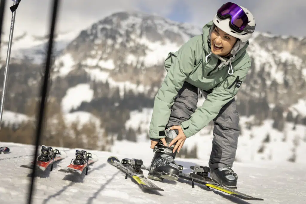 Ihr seht eine lachende Frau, die sich zu ihren Füßen beugt und die Schnalle an ihrem Skischuh schließt. Neben ihr liegen zwei Paar Ski, im Hintergrund verschneite Landschaft.