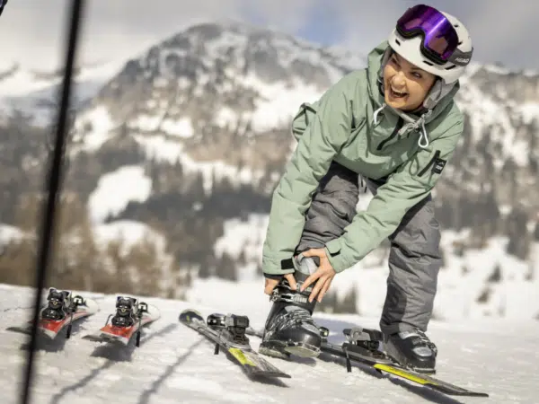 Ihr seht eine lachende Frau, die sich zu ihren Füßen beugt und die Schnalle an ihrem Skischuh schließt. Neben ihr liegen zwei Paar Ski, im Hintergrund verschneite Landschaft.