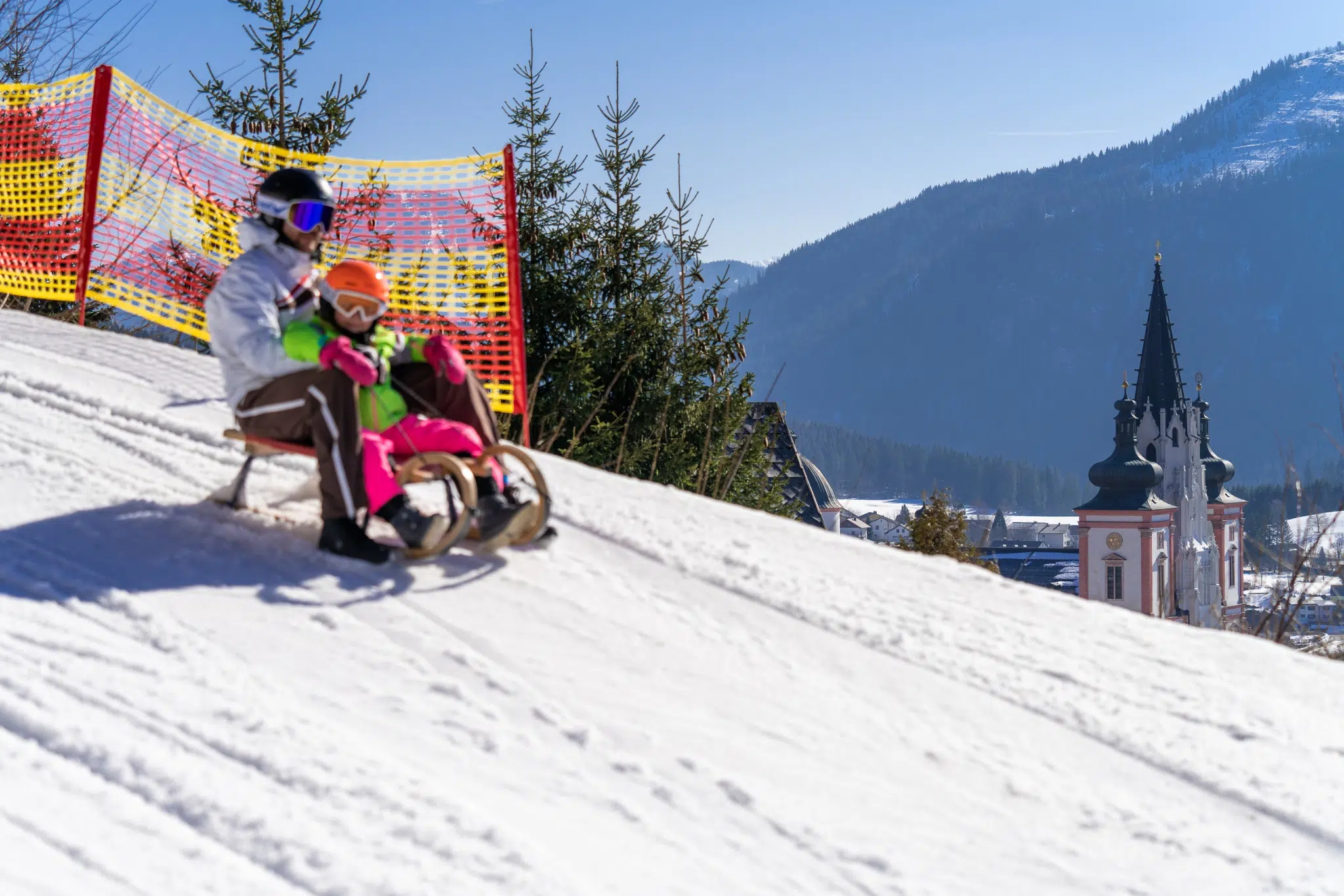 Ihr seht einen Vater mit Kind beim Rodeln auf der Mariazeller Bürgeralpe. JUFA Hotels bietet erholsamen Familienurlaub und einen unvergesslichen Winterurlaub.