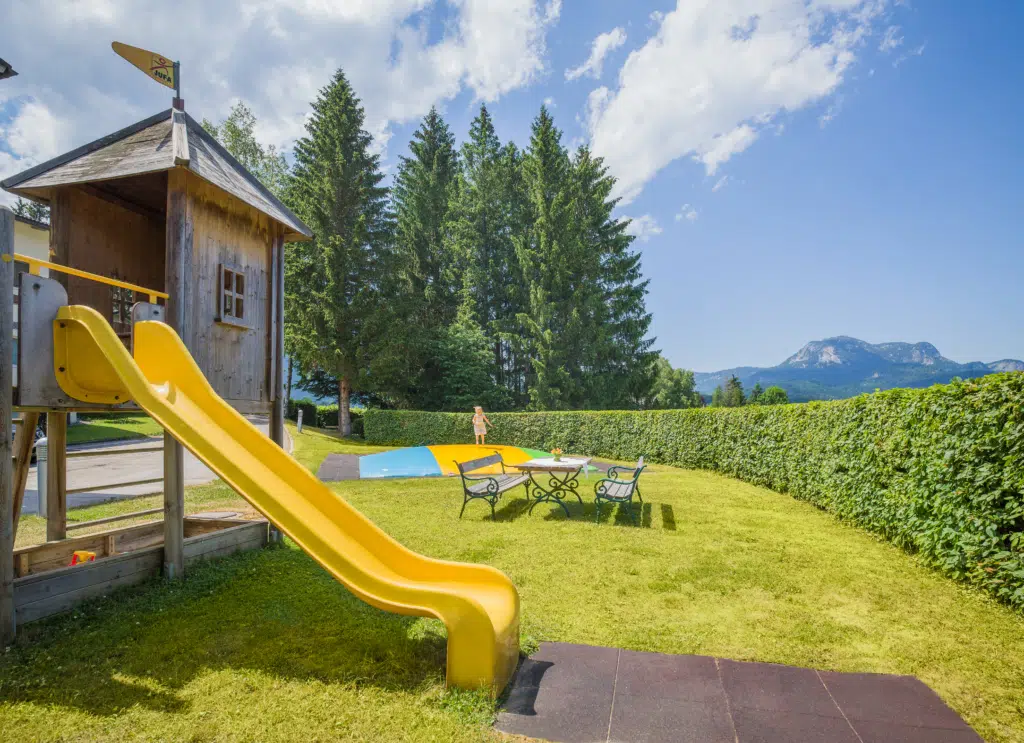 Ihr seht den Spielplatz im Garten mit Rutsche, Luftkissen, Schaukel und Kletterhaus vom JUFA Hotel Bad Aussee***. Der Ort für tollen Sommerurlaub an schönen Seen für die ganze Familie.