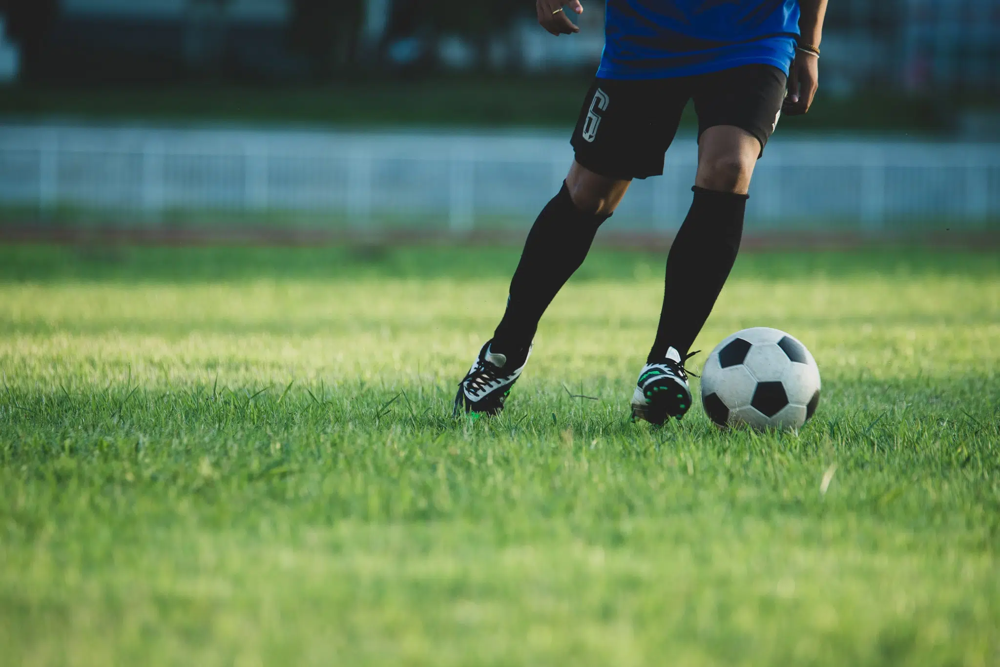 Ihr seht die Beine eines Fußballspielers, der auf einem grünen Rasen einen Fußball kickt.