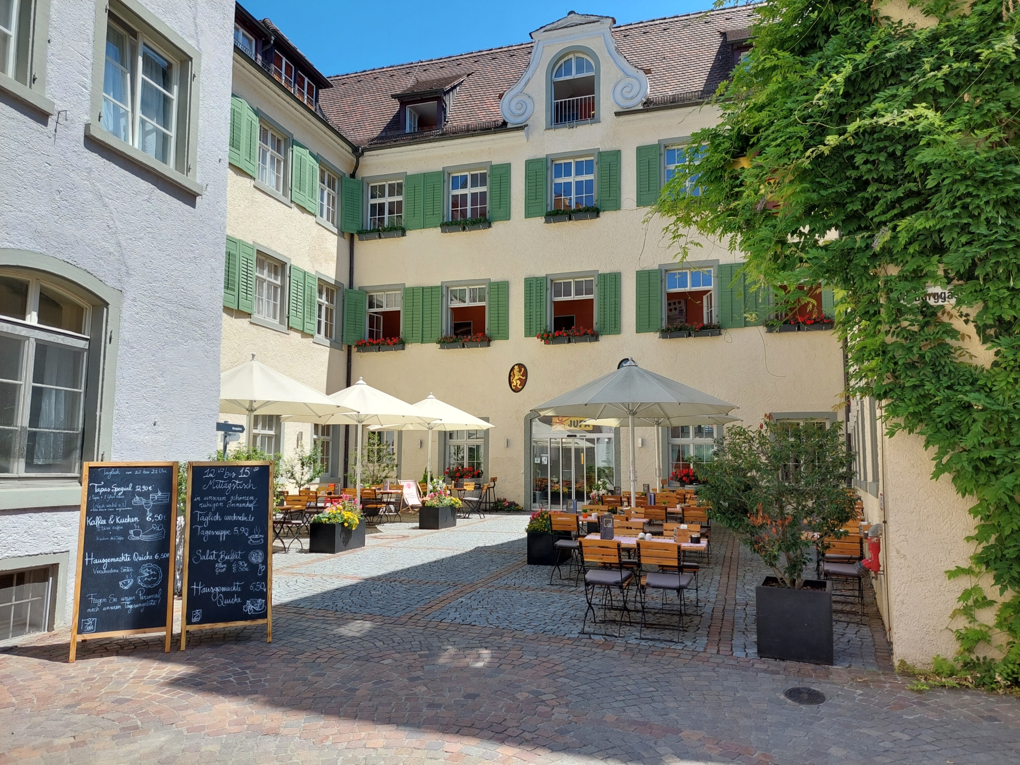 Ihr seht die Sonnenterrasse mit Tischen, Stühlen und Sonnenschirmen im Innenhof im JUFA Hotel Meersburg am Bodensee in Baden-Württemberg in Deutschland.