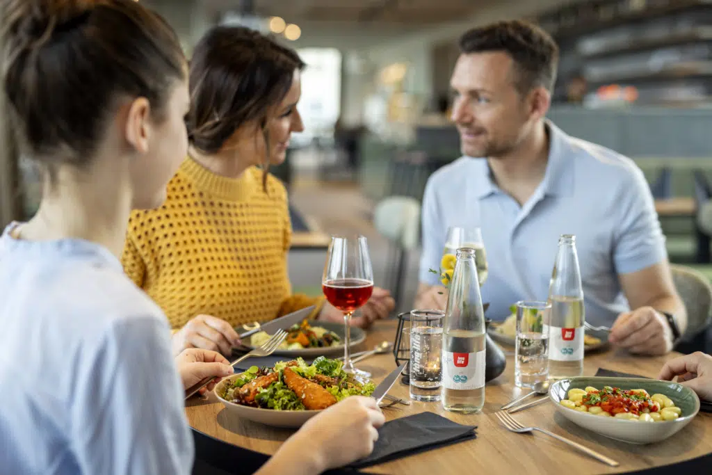 Ihr seht eine Familie an einem Tisch im Café & Restaurant Wandelhalle, die gerade regionale Speisen und Getränke aus dem Steirischen Thermen- & Vulkanland genießt.