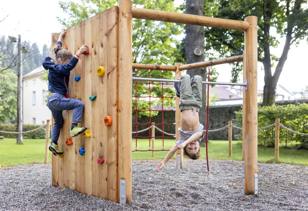 Ihr seht einen Jungen, der am Spielplatz auf der Kletterstange kopfüber hängt und seine Arme schwingt. Daneben klettert die Schwester die Kletterwand hoch. JUFA Hotels bietet kinderfreundlichen und erlebnisreichen Urlaub für die ganze Familie.