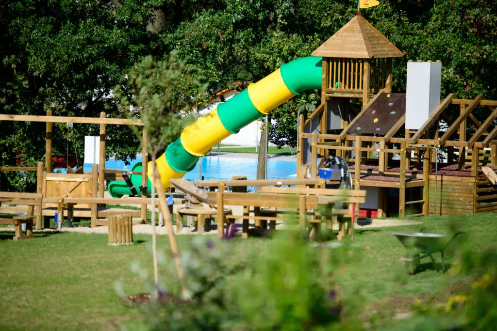 Ihr seht den Spielplatz mit Holzgeräten und einer Rutsche und Bäumen im Hintergrund beim JUFA Hotel Neutal Landerlebnis. Die Zeit spielend und abwechslungsreich verbringen im JUFA Hotel Neutal - Landerlebnis. Der Ort für erlebnisreichen Natururlaub für die ganze Familie.