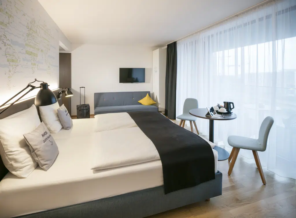 Ihr seht ein Doppelbett und eine Couch in einer Suite des JUFA Hotels Hamburg HafenCity****. Der Ort für erlebnisreichen Städtetrip für die ganze Familie und der ideale Platz für Ihr Seminar.