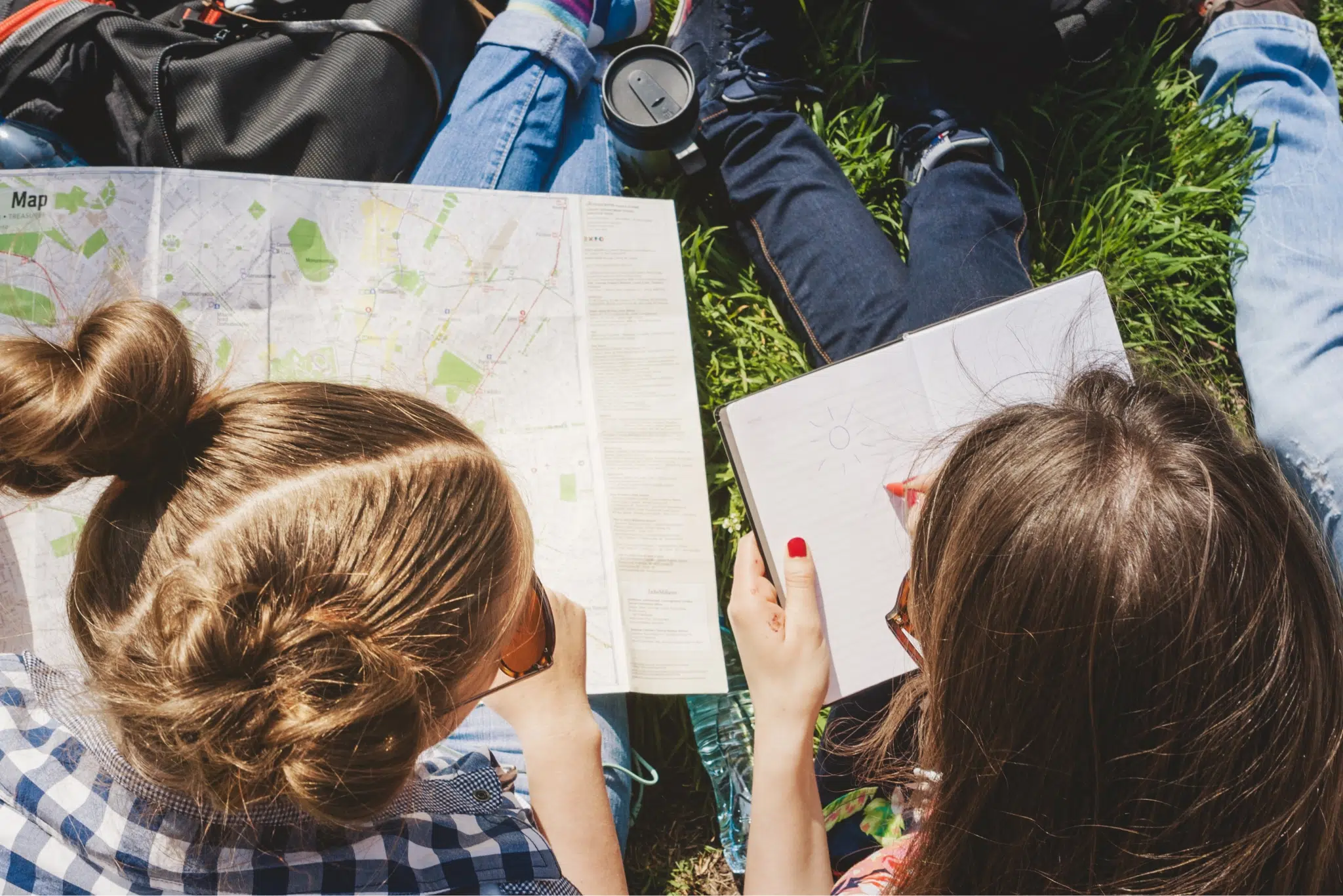 Ihr seht zwei Teenager-Mädchen mit einem Stadtplan und einem Notizbuch, die in einer Wiese sitzen.