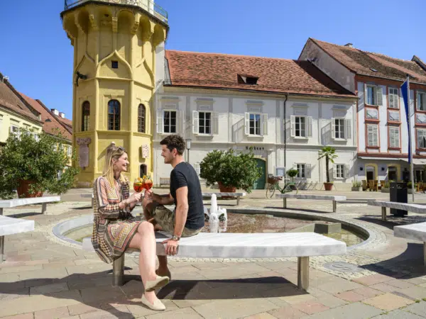 Ihr seht einen Mann und eine Frau beim Aperitif auf einer Bank in der Altstadt von Bad Radkersburg im Sommer.