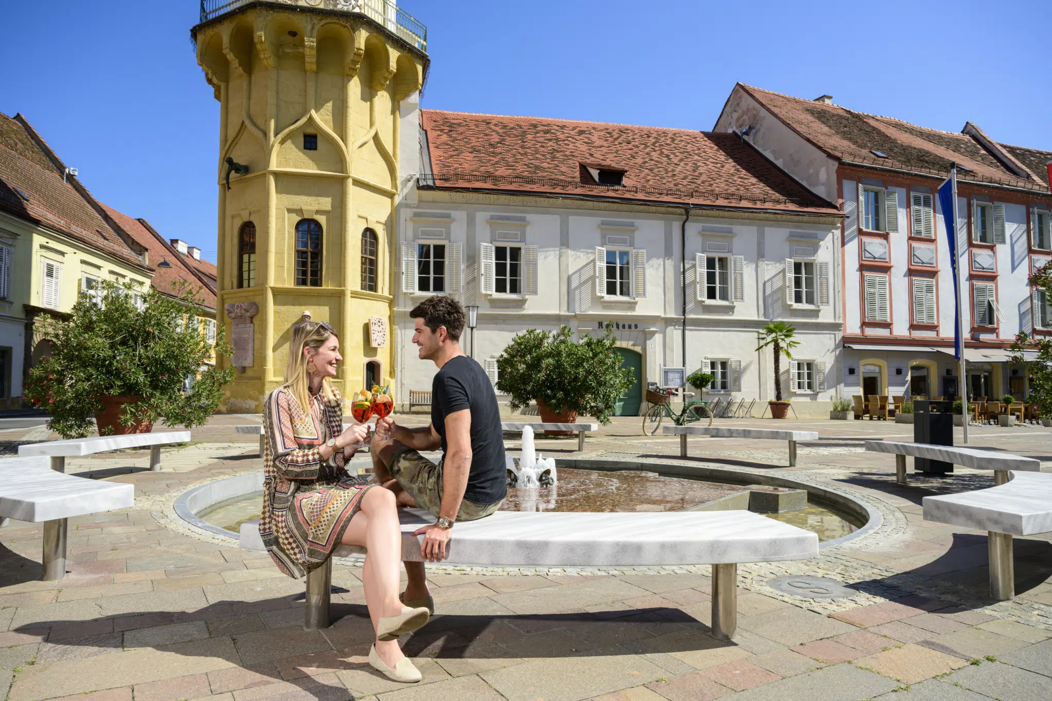 Ihr seht einen Mann und eine Frau beim Aperitif auf einer Bank in der Altstadt von Bad Radkersburg im Sommer.