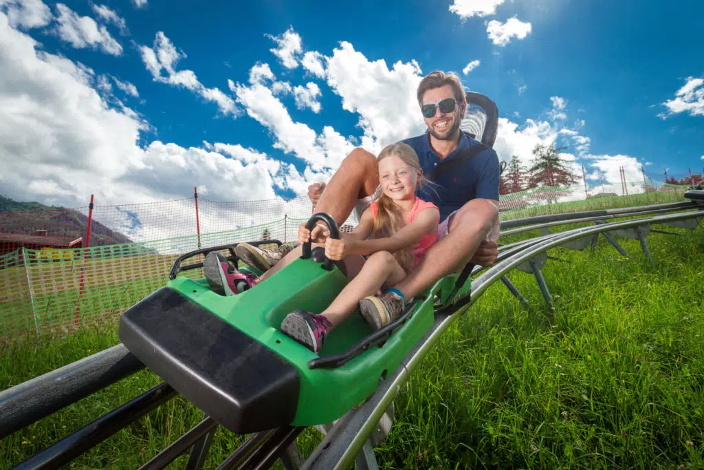 Ihr seht einen Vater und seine Tochter mit einem Maisiflitzer an einem Sommertag ins Tal fahren.