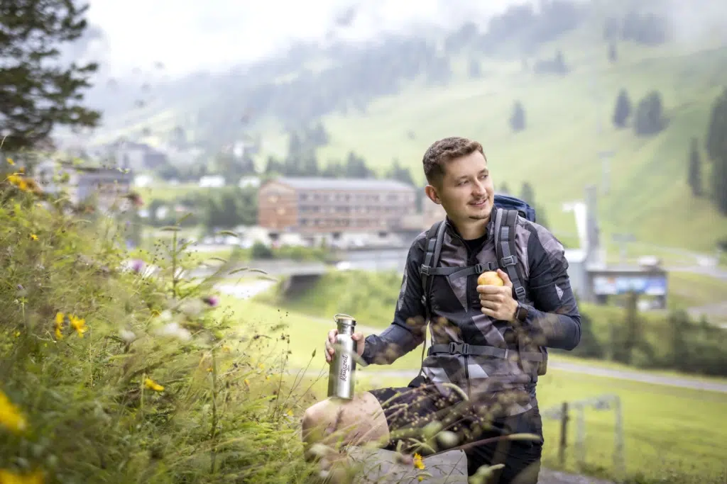 Sie sehen einen Wanderer vor dem JUFA Hotel Malbun mit Jause und der Rauszeit-Thermosflasche