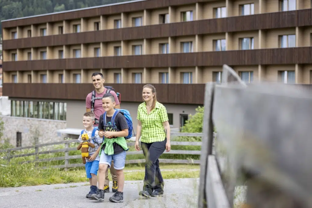 Ihr seht eine Familie, die zum Wandern aufbricht mit dem JUFA Hotel Savognin***s im Hintergrund.