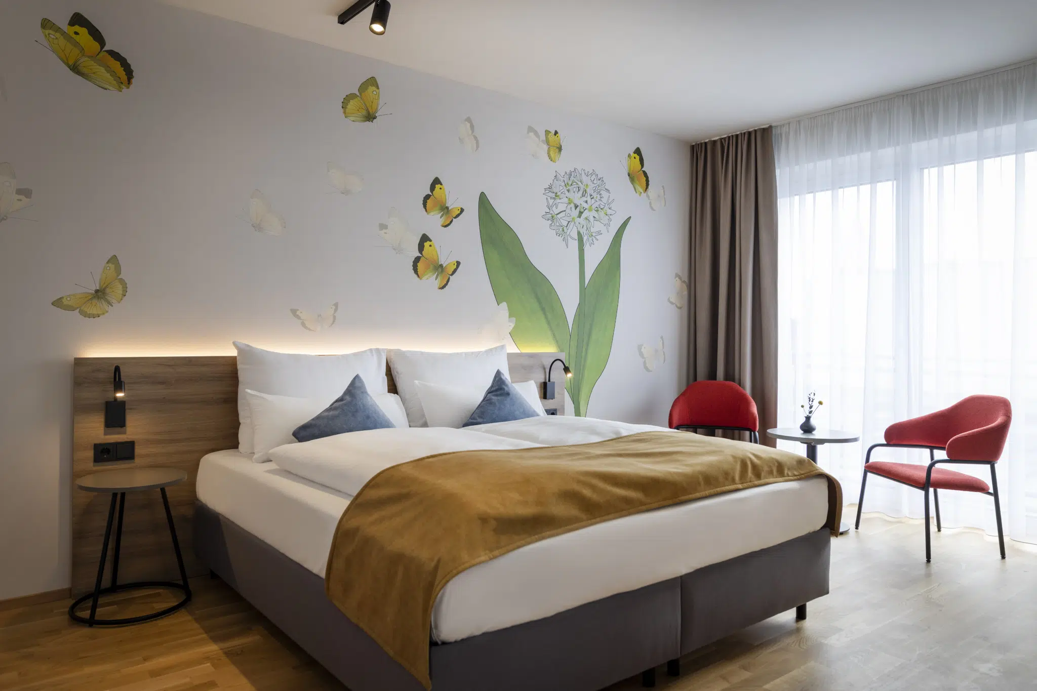 Sie sehen ein Doppelzimmer für zwei Personen im JUFA Hotel Bad Radkersburg mit Balkon, komfortablem Boxpringbett, modernem Möbiliar, gemütlicher Sitzecke mit Tisch und kreativer Wanddeko.