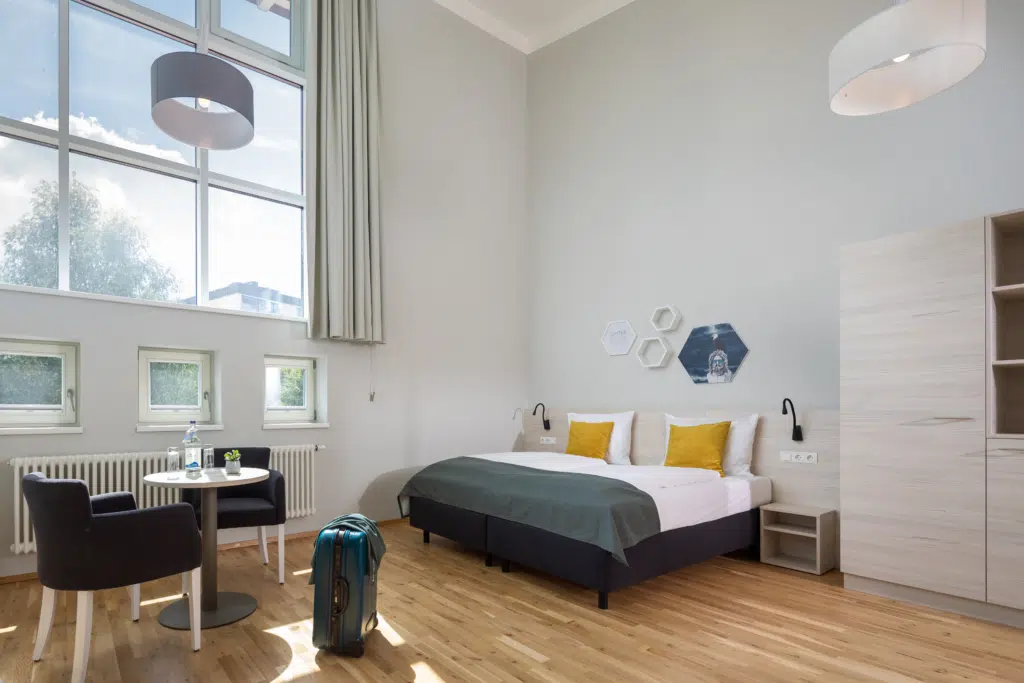 Ihr seht das Doppelbett im Doppelzimmer mit Fenster mit Sitzecke. JUFA Hotels bietet kinderfreundlichen und erlebnisreichen Urlaub für die ganze Familie.