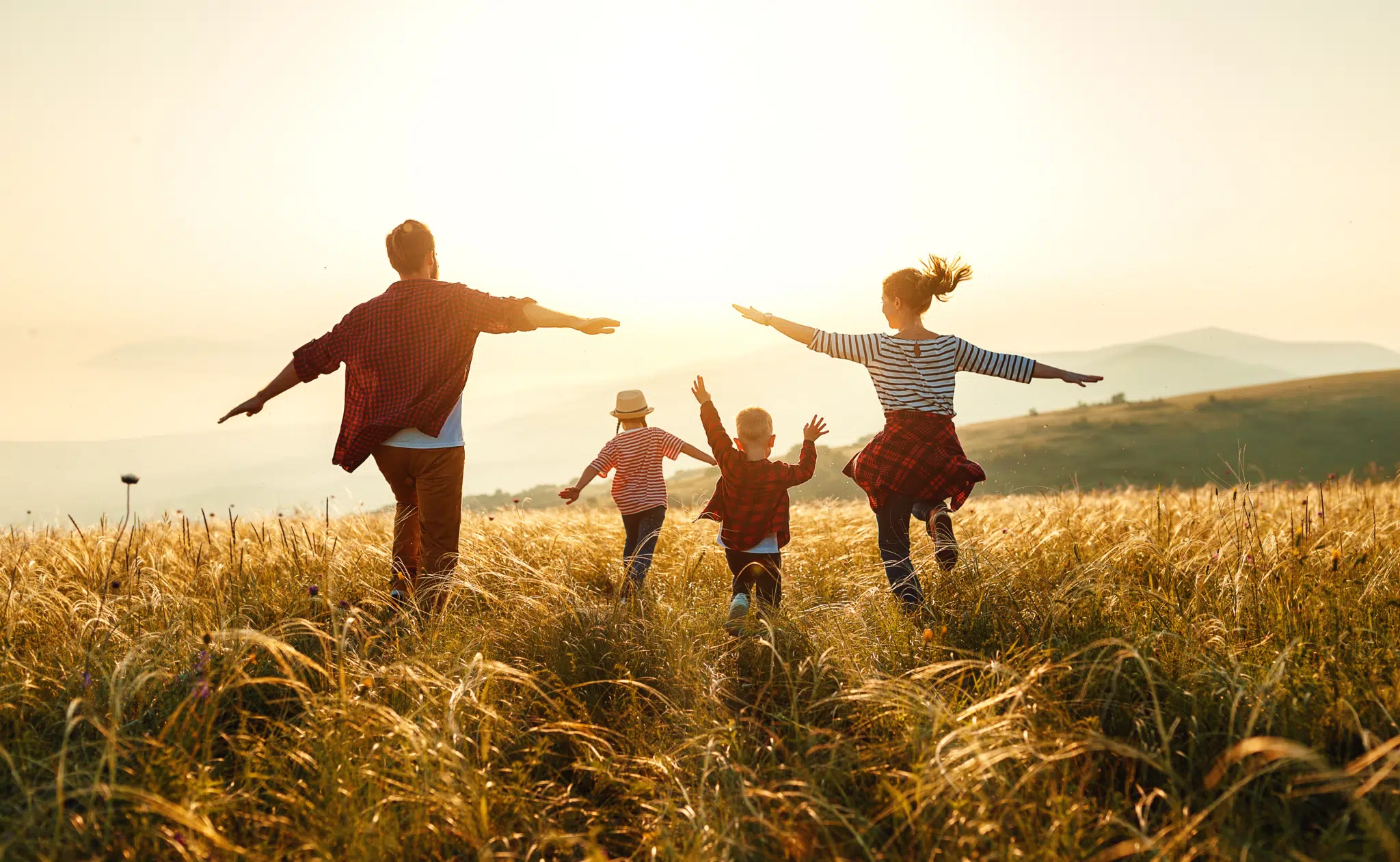 Sie sehen eine Familie mit Mutter, Vater und 2 Kindern, die mit ausgestreckten Armen über ein Getreidefeld laufen, im Hintergrund ist ein Sonnenuntergang zu sehen.