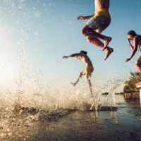 Sie sehen mehrere Personen in der sommerlichen Abenddämmerung, die gerade von einer Plattform ins Wasser springen in der Nähe der JUFA Hotels.