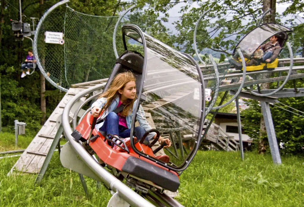 Ihr seht ein Mädchen, das im Alpine Coaster am Erlebnisberg Wurbauerkogel nach unten fährt. Hinter ihr fährt ein Mann mit seinem Kind.