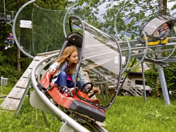 Ihr seht ein Mädchen, das im Alpine Coaster am Erlebnisberg Wurbauerkogel nach unten fährt. Hinter ihr fährt ein Mann mit seinem Kind.