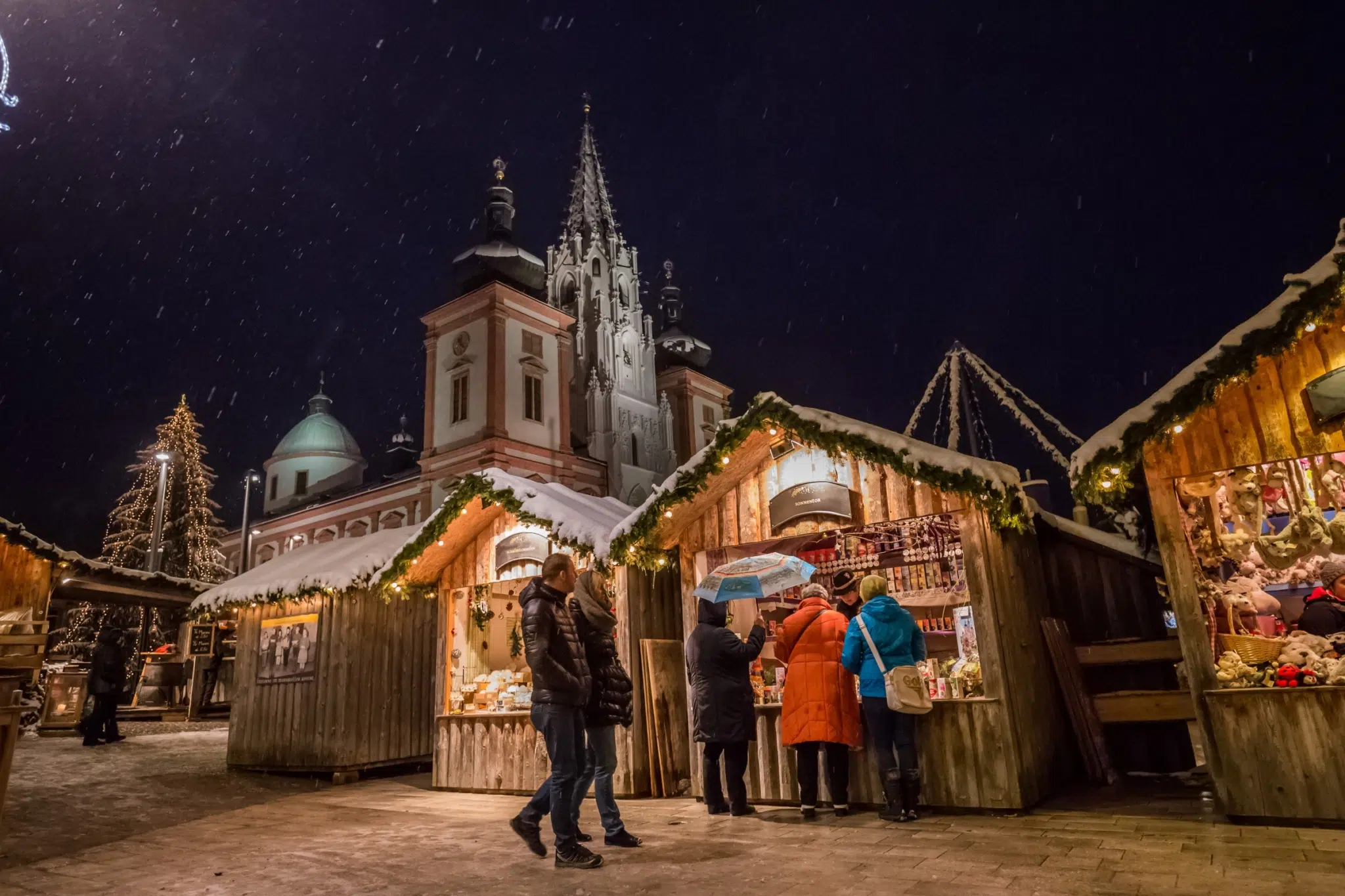 Ihr seht Stände am Adventmarkt im verschneiten Mariazell mit der Basilika im Hintergrund.