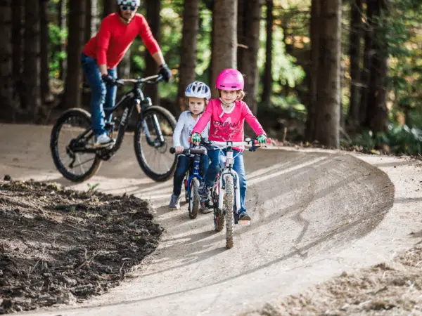 Ihr seht einen Vater mit zwei Kindern beim Mountainbiken im Mini-Bikepark beim JUFA Hotel Annaberg. Der Ort für erholsamen Familienurlaub und einen unvergesslichen Winter- und Wanderurlaub.