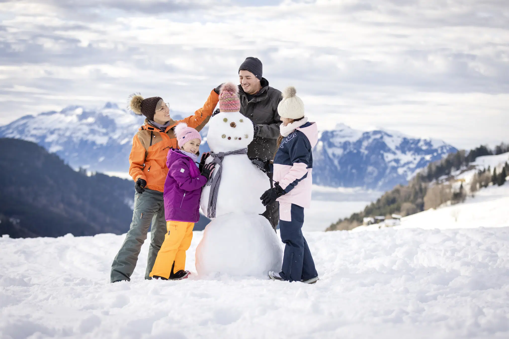 Ihr seht eine Familie, die einem Schneemann eine Mütze aufsetzt. Das JUFA Hotel Laterns - Klangholzhus ist der ideale Ort für einen abwechslungsreichen Familien-Winterurlaub und unvergessliche Tage im Schnee.