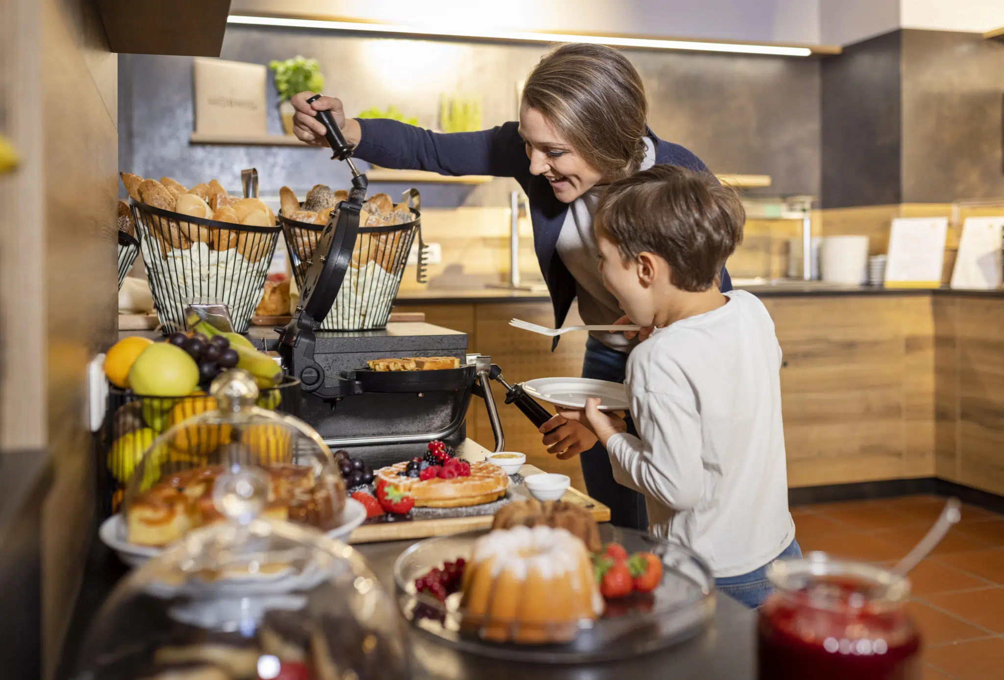 Sie sehen wie die Mutter ihrem Sohn hilft Waffeln beim Frühstücksbuffet zuzubereiten. JUFA Hotels bietet erholsamen Familienurlaub und einen unvergesslichen Winter- und Wanderurlaub.