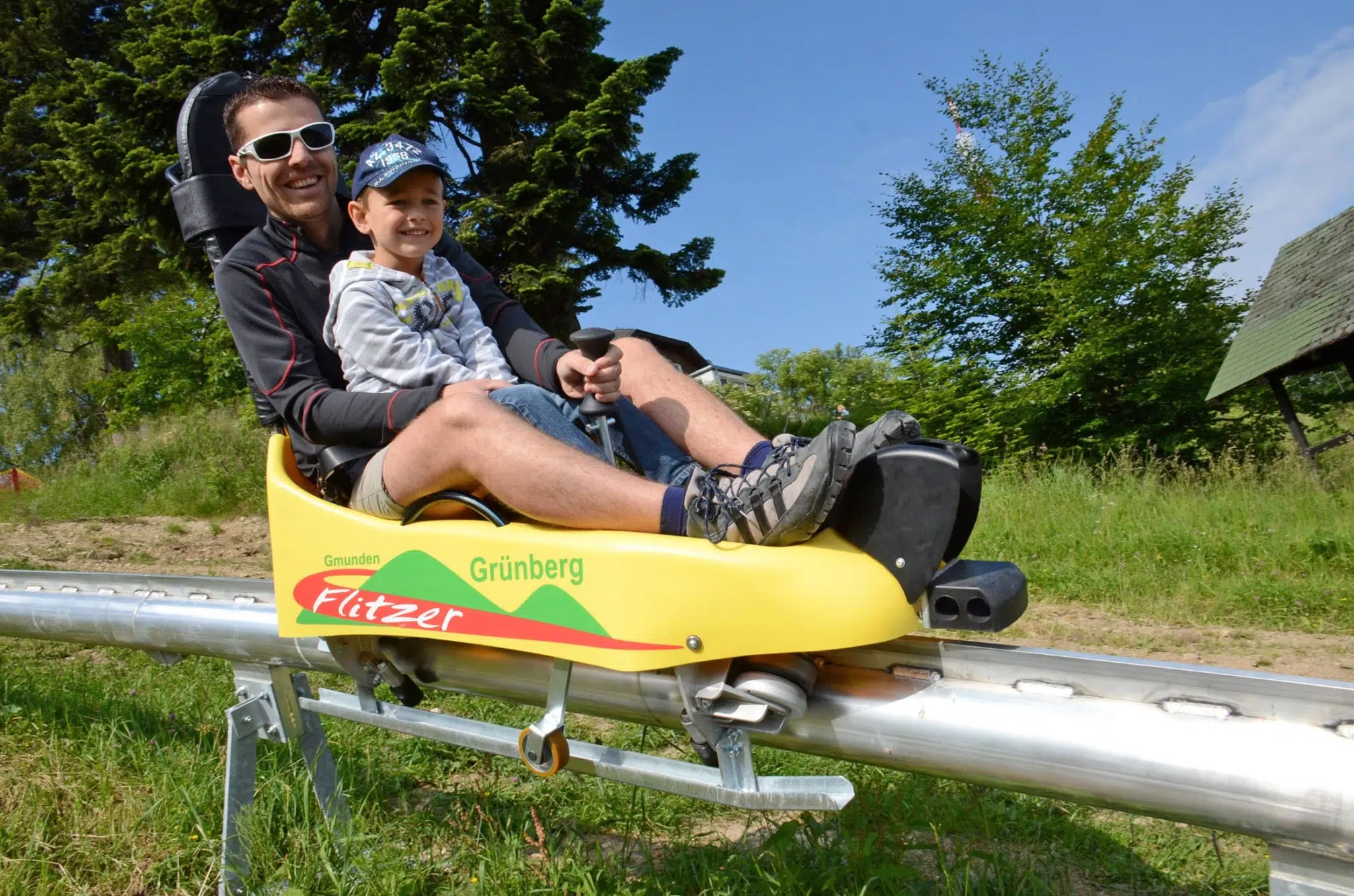 Vater und Sohn sausen mit einer Sommerrodel des Grünberg Flitzers im Salzkammergut ins Tal. JUFA Hotels bietet kinderfreundlichen und erlebnisreichen Urlaub für die ganze Familie.