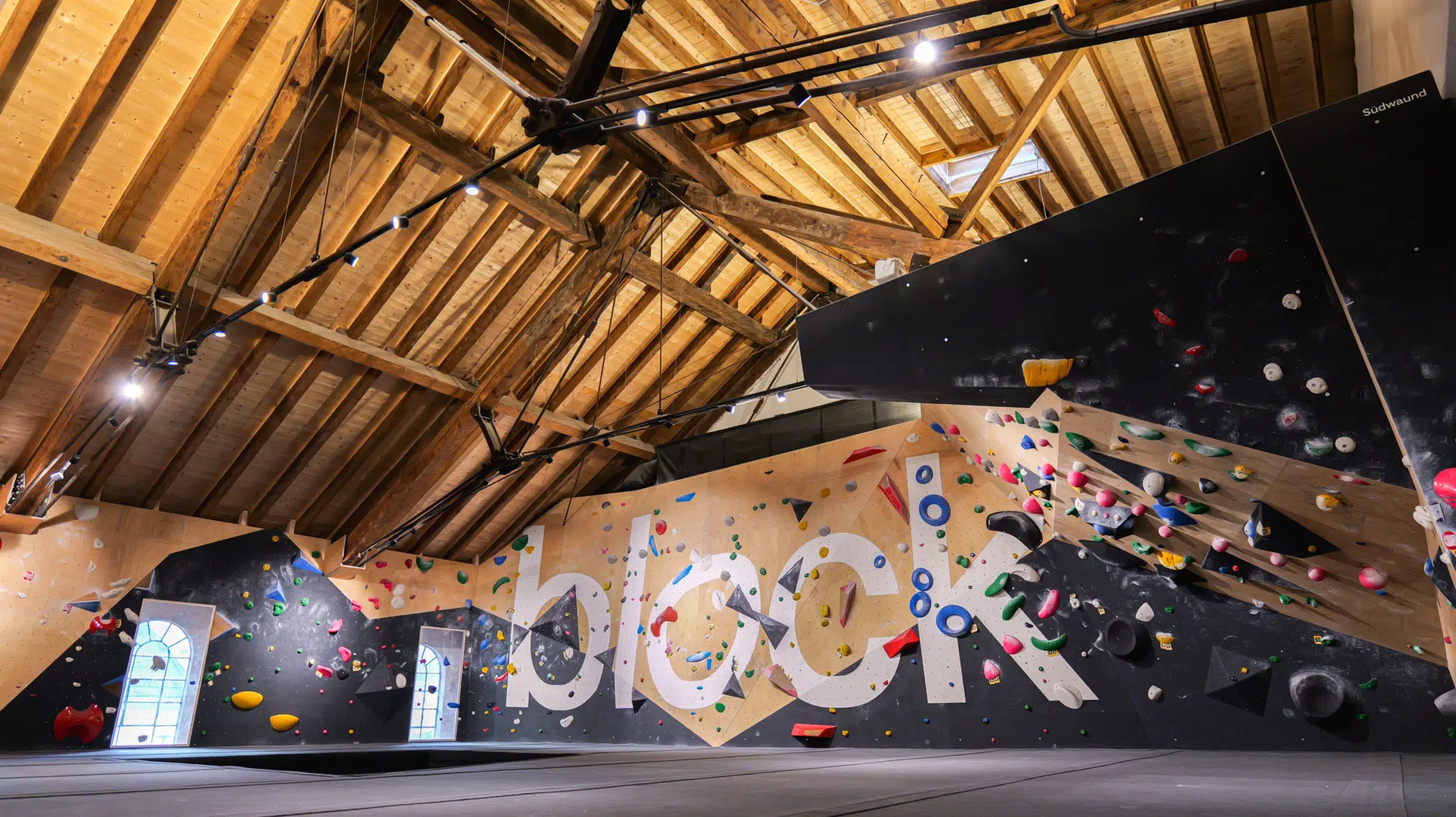 Ihr seht einen Teil der stoablock Boulderhalle mit Boulderwand, Matten und Holzgiebel-Dachstuhl.