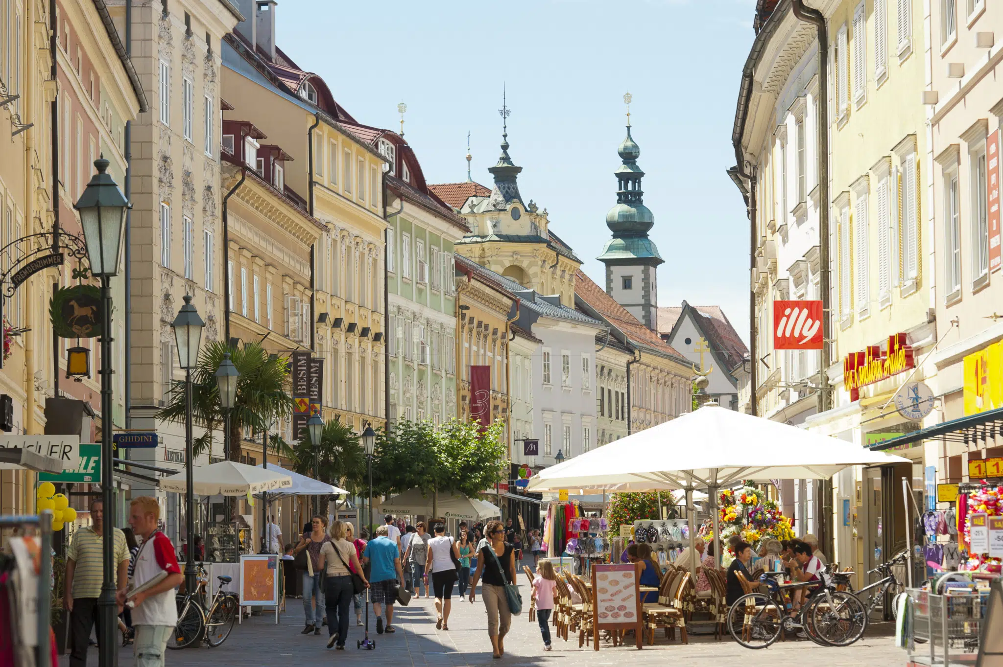 Ihr seht eine Straße in der Altstadt von Klagenfurt in Kärnten mit Türmchen, historischen Häuserfassaden und Gastgärten.