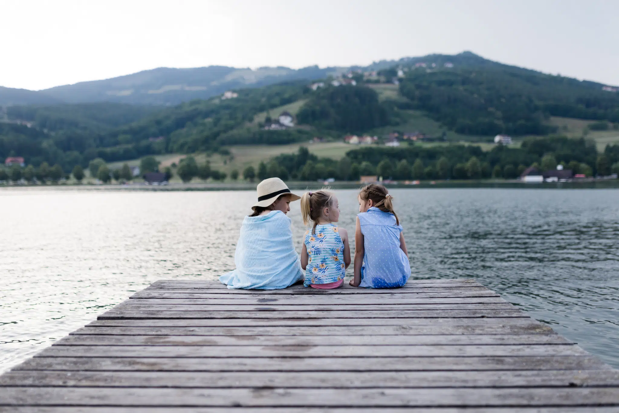 Sie sehen drei Mädchen auf einem Steg mit Blick auf den Stubenbergsee im Sommer. JUFA Hotels bietet tollen Sommerurlaub an schönen Seen für die ganze Familie.