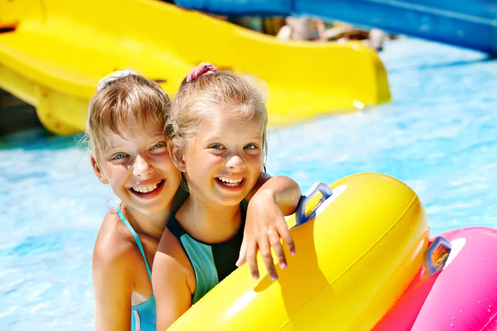 Sie sehen zwei lachende blonde Mädchen mit Schwimmreifen im Freibad mit Rutsche. JUFA Hotels bietet erholsamen Thermen- und Badespass für die ganze Familie.