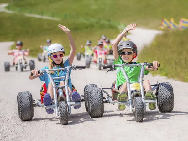 Ihr seht Kinder auf Mountaincarts auf einem Kiesweg auf der Mariazeller Gemeindealpe in der Nähe von JUFA Hotels fahren. Der Ort für erholsamen Familienurlaub und einen unvergesslichen Winter- und Wanderurlaub.