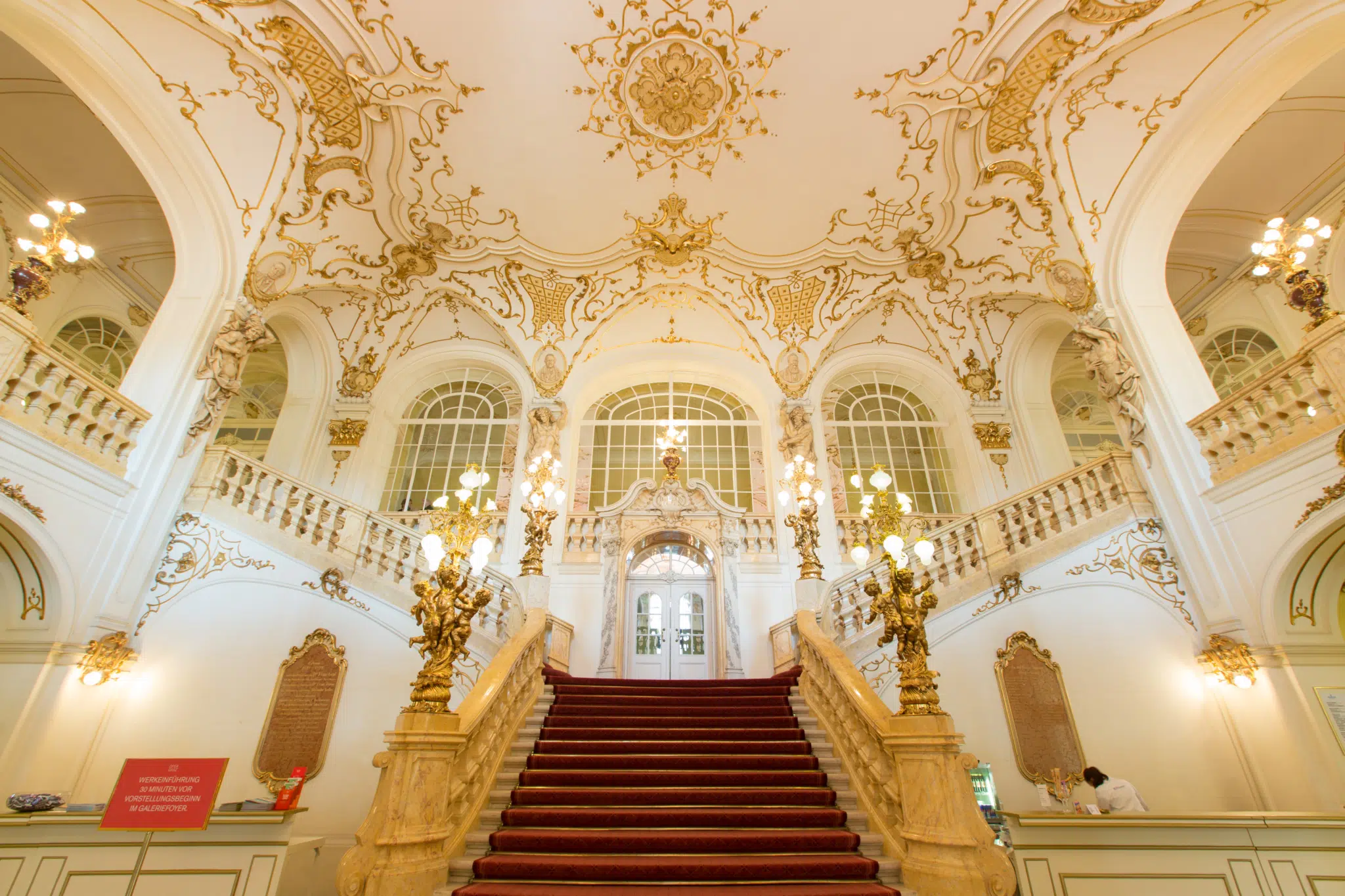 Ihr seht das prachtvolle Foyer der Oper Graz mit Treppe, rotem Teppich und prunkvoller Goldverzierung.