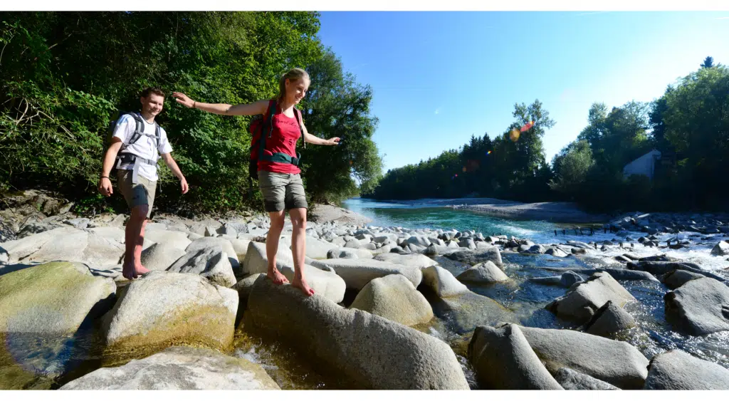 Ein junges Paar balanciert im Sommer über große Steine im Flussbett des Almflusses in Oberösterreich. JUFA Hotels bietet Ihnen den Ort für erlebnisreichen Natururlaub für die ganze Familie.