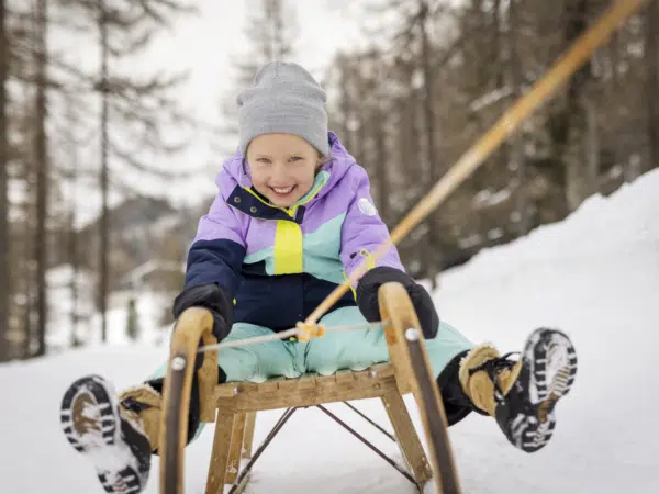 Ihr seht ein Mädchen in Skikleidung, das lachend auf einer Rodel in einem Winterwald gezogen wird.