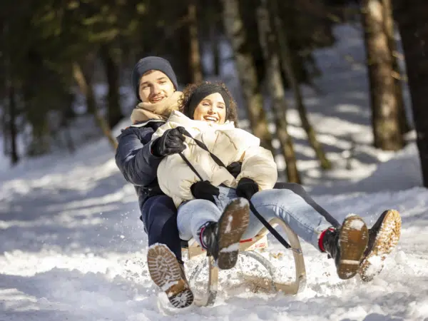 Ihr seht ein junges Paar beim Schlittenfahren durch den Wald. Das JUFA Hotel Laterns - Klangholzhus ist ein idealer Ort für einen unvergesslichen Winter- und Familienurlaub und erholsame Tage für die ganze Familie.