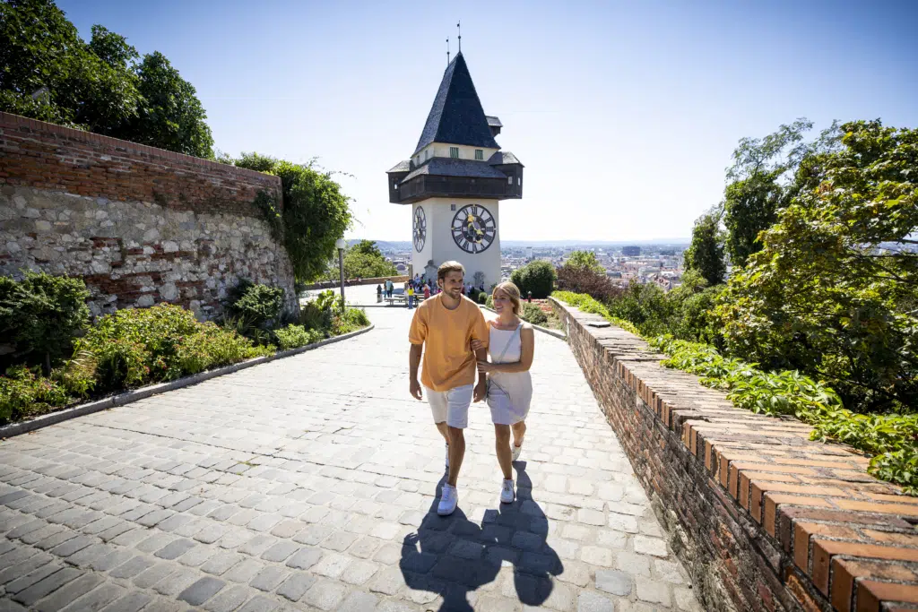 Sie sehen einen jungen Mann und eine junge Frau in sommerlicher Bekleidung im Sonnenschein vor dem Uhrturm am Grazer Schloßberg.