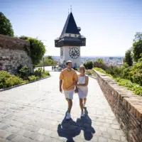 Sie sehen einen jungen Mann und eine junge Frau in sommerlicher Bekleidung im Sonnenschein vor dem Uhrturm am Grazer Schloßberg.