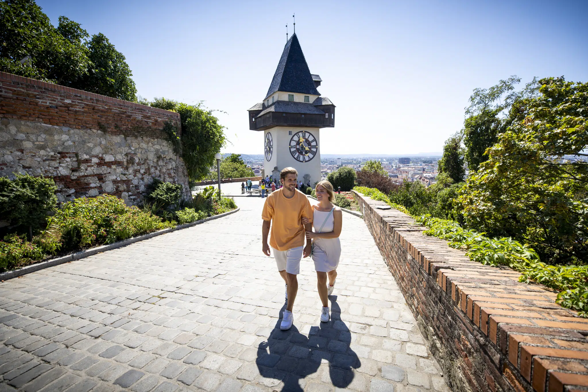 Sie sehen einen jungen Mann und eine junge Frau in sommerlicher Bekleidung im Sonnenschein vor dem Uhrturm am Grazer Schloßberg.