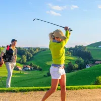 Sie sehen eine Frau und einen Mann beim Golfspielen, gerade beim Abschlag im Hintergrund mit grünen Hügeln und Sandbunker.